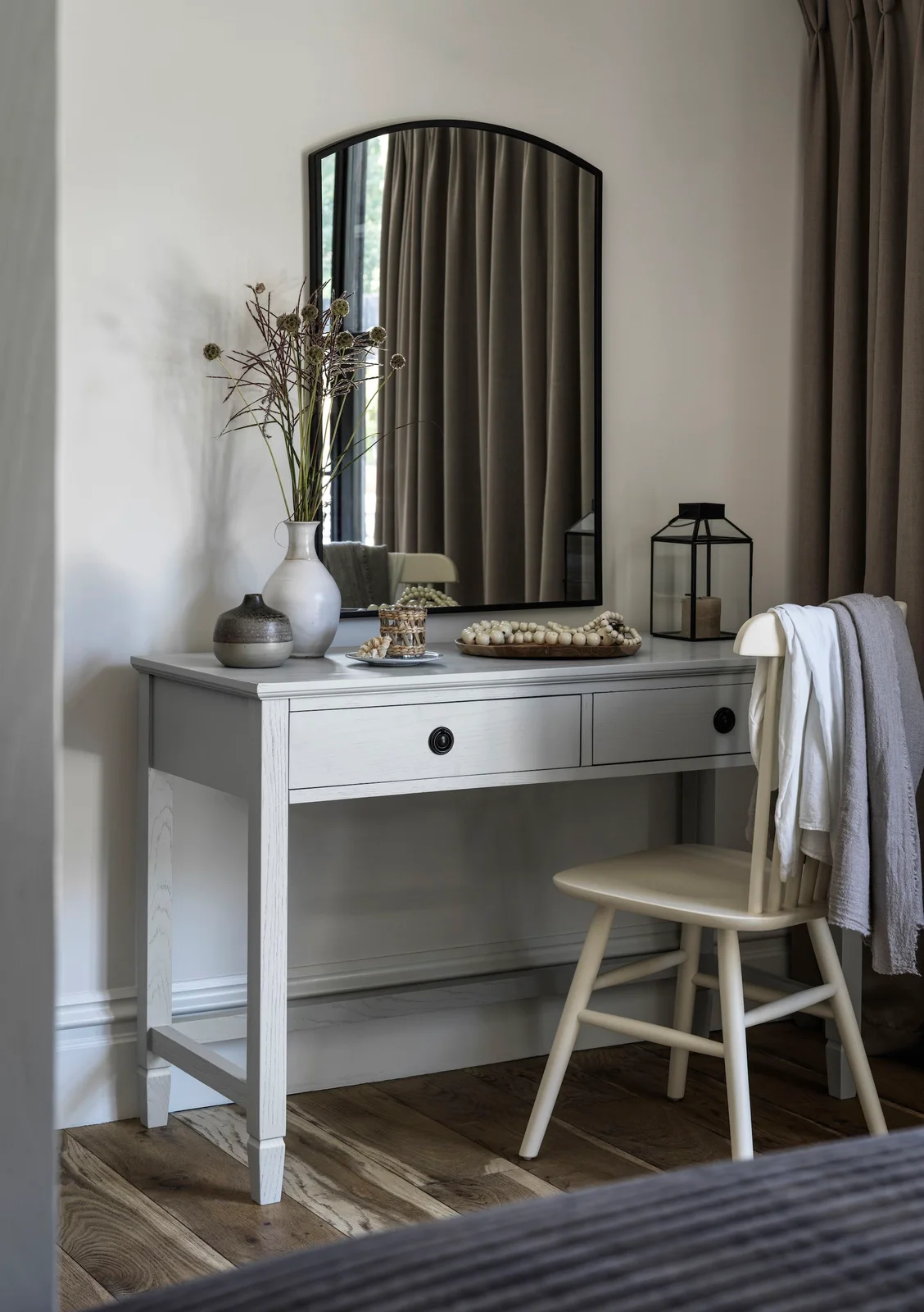 A pale gray two-drawer dressing table sits below a tall arched mirror beside a cream chair