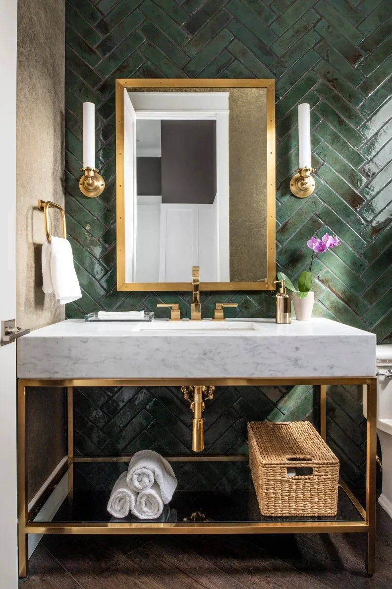 Dark green herringbone tiles stand behind a pale basin counter on a warm-metal console, with a rectangular mirror, two slim wall lights and a woven storage basket.