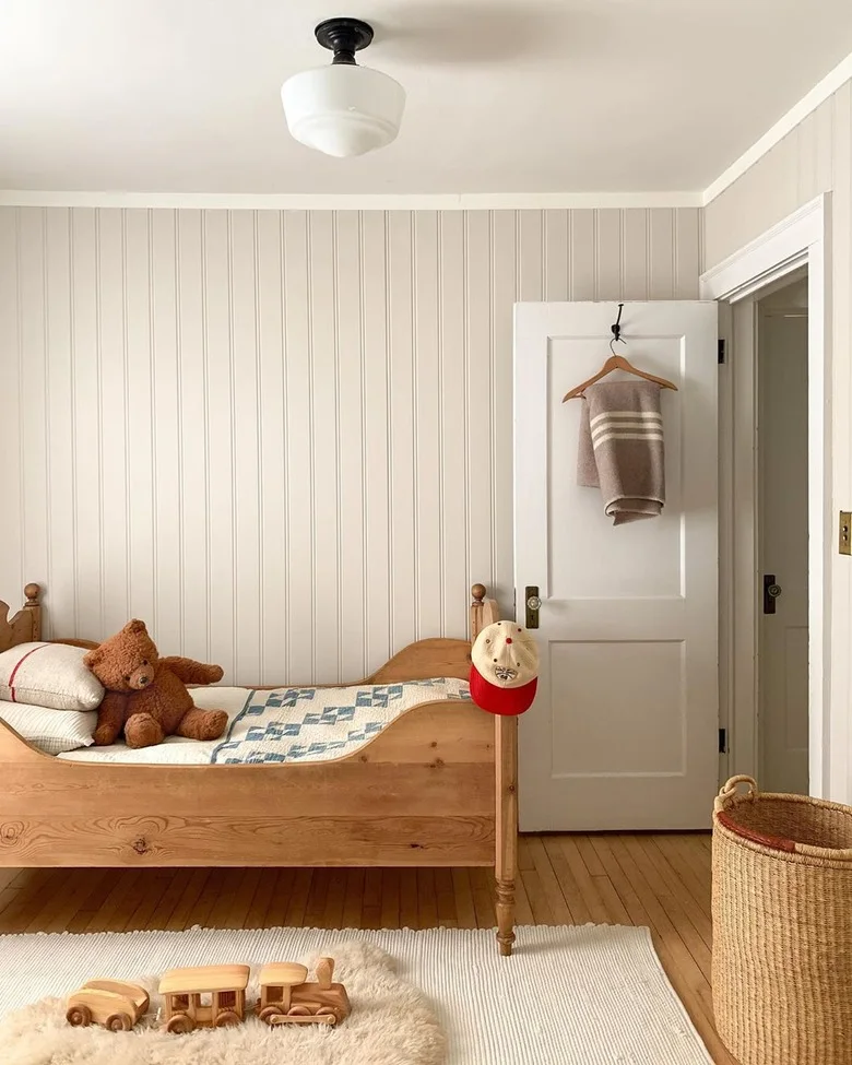 A wood single bed with a checked cover and teddy bear stands against pale vertical paneling beside a woven basket and a light rug holding small wood toys