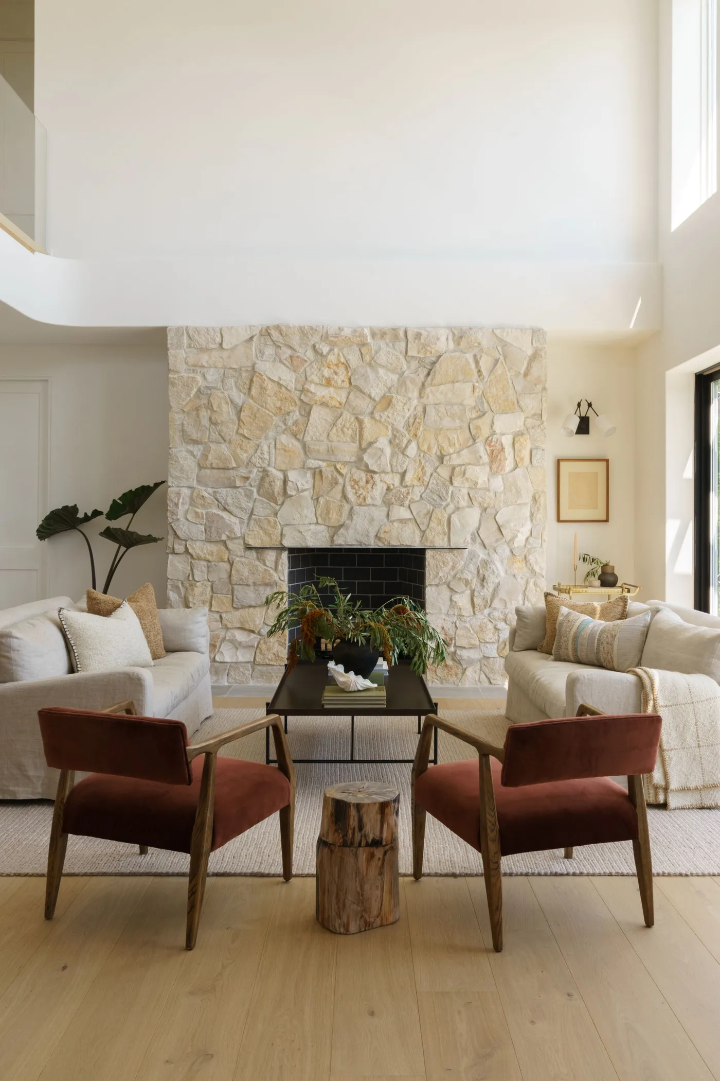 Tall pale irregular stone fireplace wall with black firebox, pale sofas and rust-colored chair