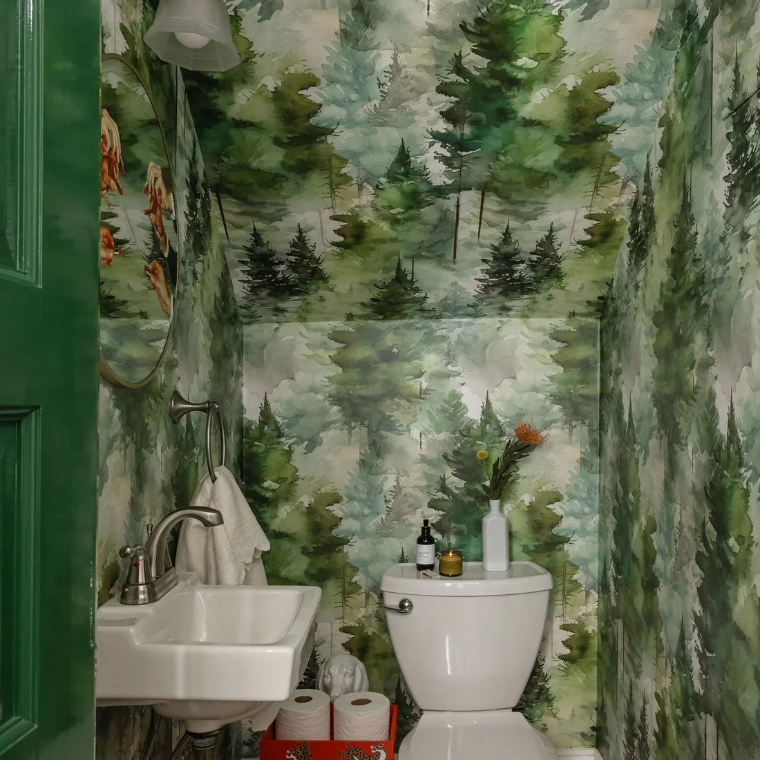 Green illustrated tree wallpaper wraps a narrow powder room around a white toilet and compact wall sink beside a green door