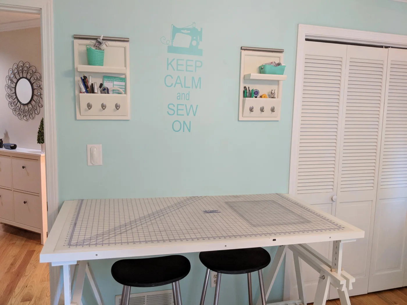 A white trestle table with a large cutting mat and two dark stools stands below wall organizers beside white closet doors.