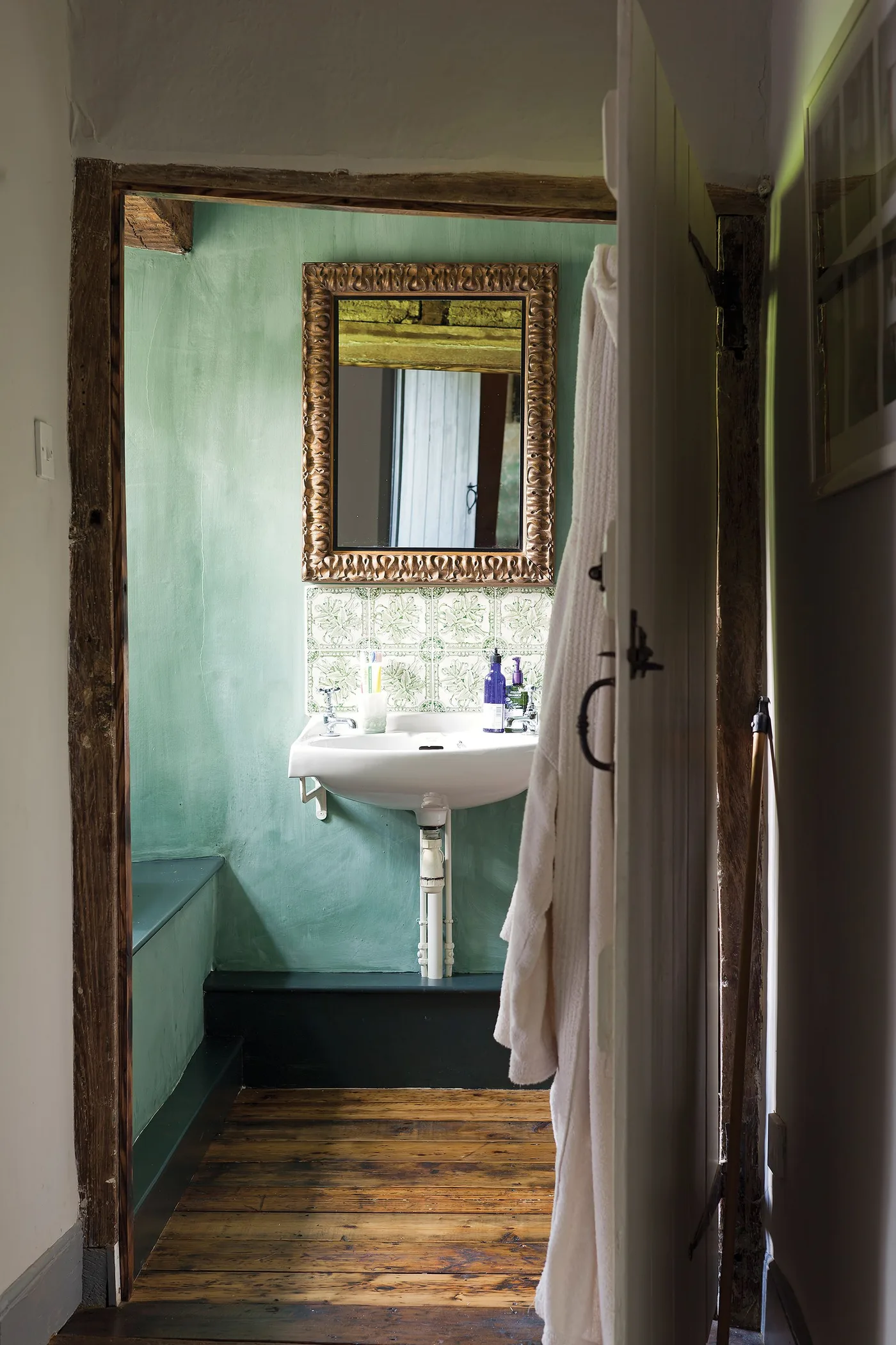 Small bathroom doorway with a wooden frame, blue-green walls, white wall-mounted basin, ornate mirror and warm wooden floor.