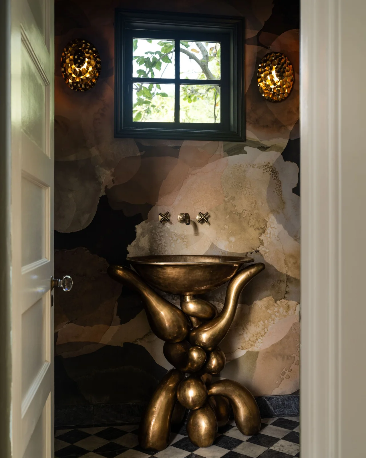 A tiny powder room has a gold-toned sculptural basin stand, large softly patterned walls, a dark-framed window, decorative wall lights and a checkerboard floor.