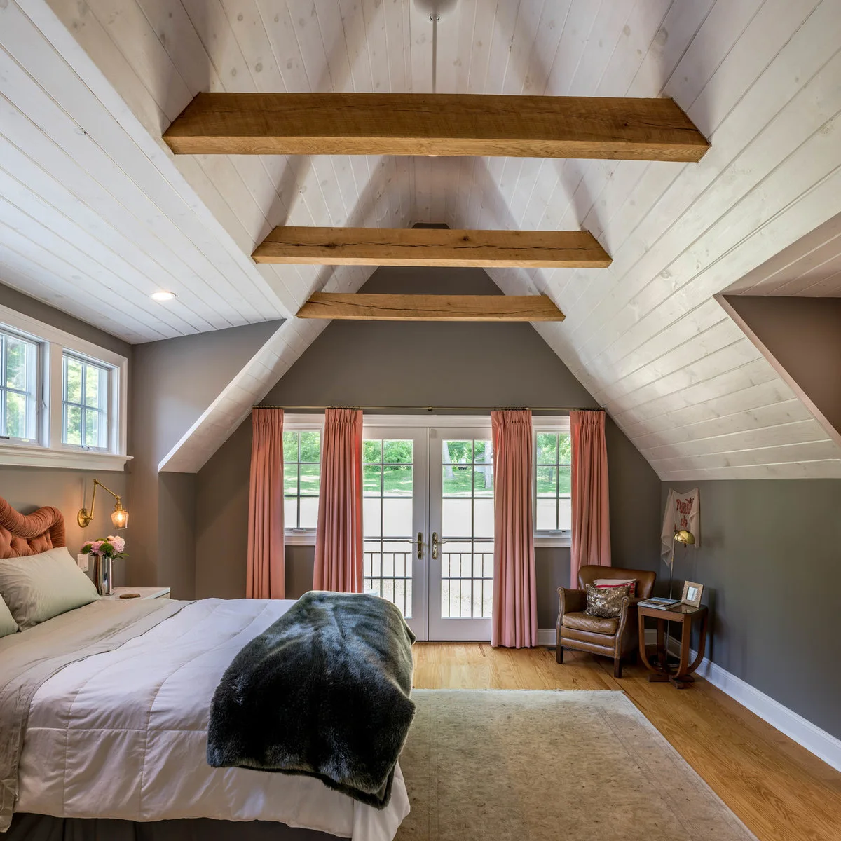 Attic bedroom with white boarded slopes, exposed brown beams, pink curtains and a brown chair beside a side table.