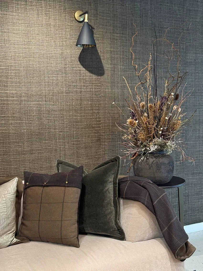 A tall arrangement of dry branches, grasses and small flower heads stands in a round dark pot beside cushions and a dark wall sconce.