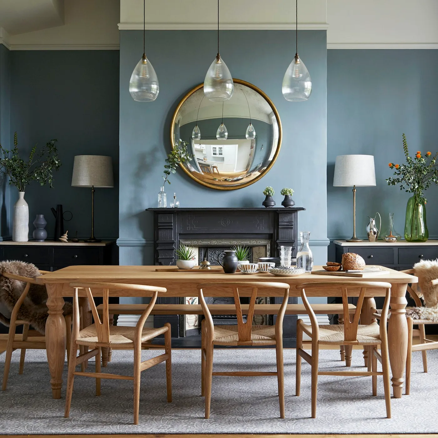Gold-toned round mirror above a black fireplace with lamps on side cabinets behind a long wood table