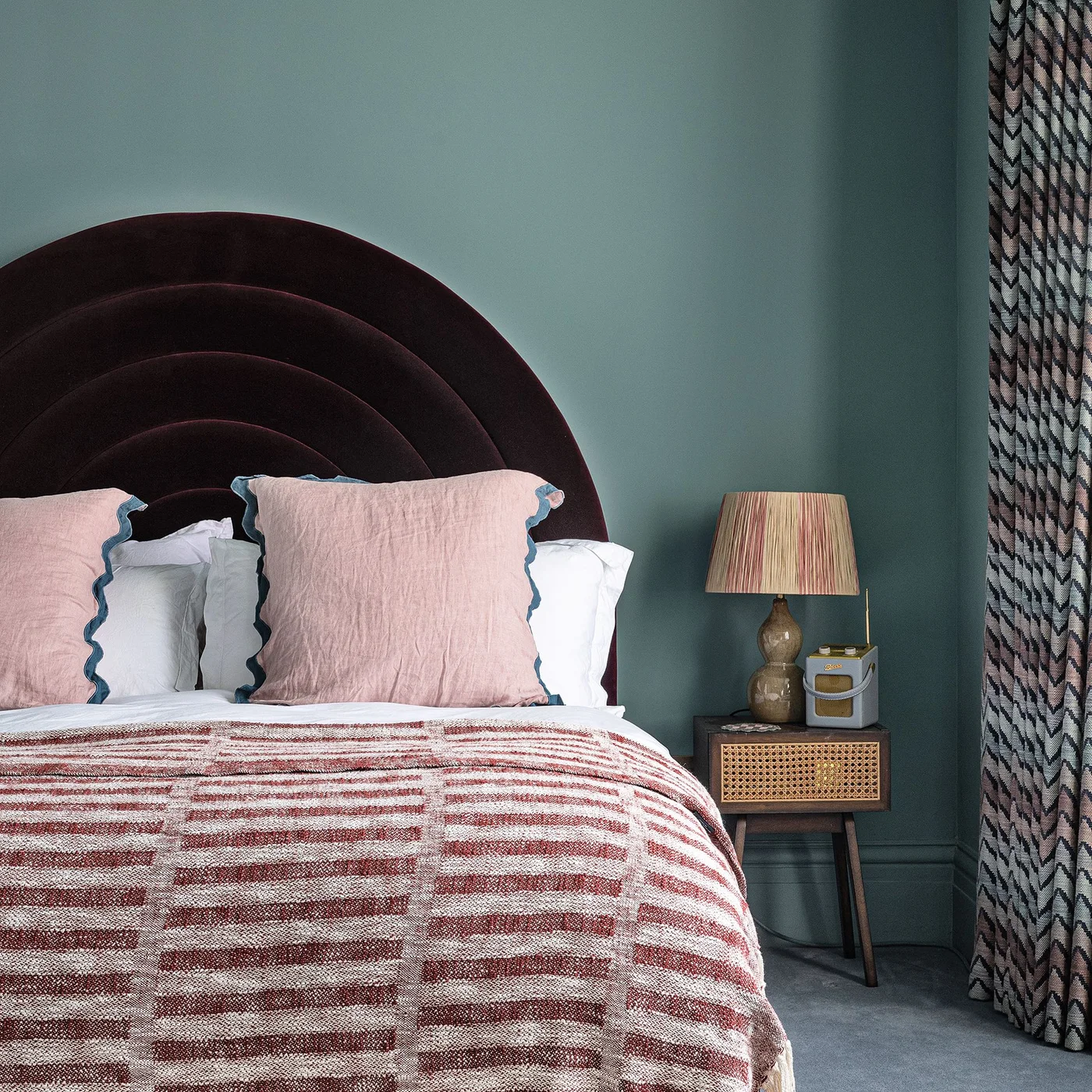 A dark wine-colored arched headboard sits against blue walls with pink scalloped pillows, a patterned throw and a small cane-front bedside cabinet.