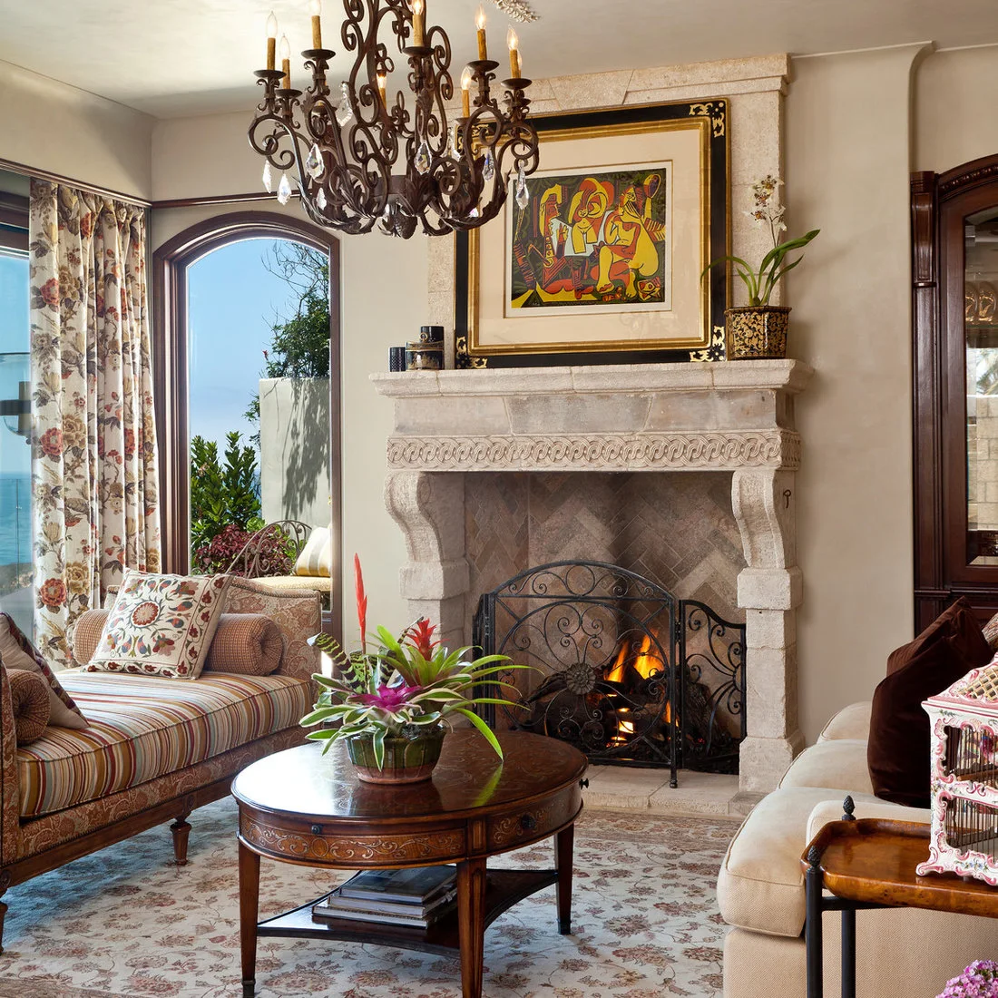 A carved pale fireplace stands beside an arched living-room door, striped and floral upholstery, a round wooden table and decorative dark chandelier