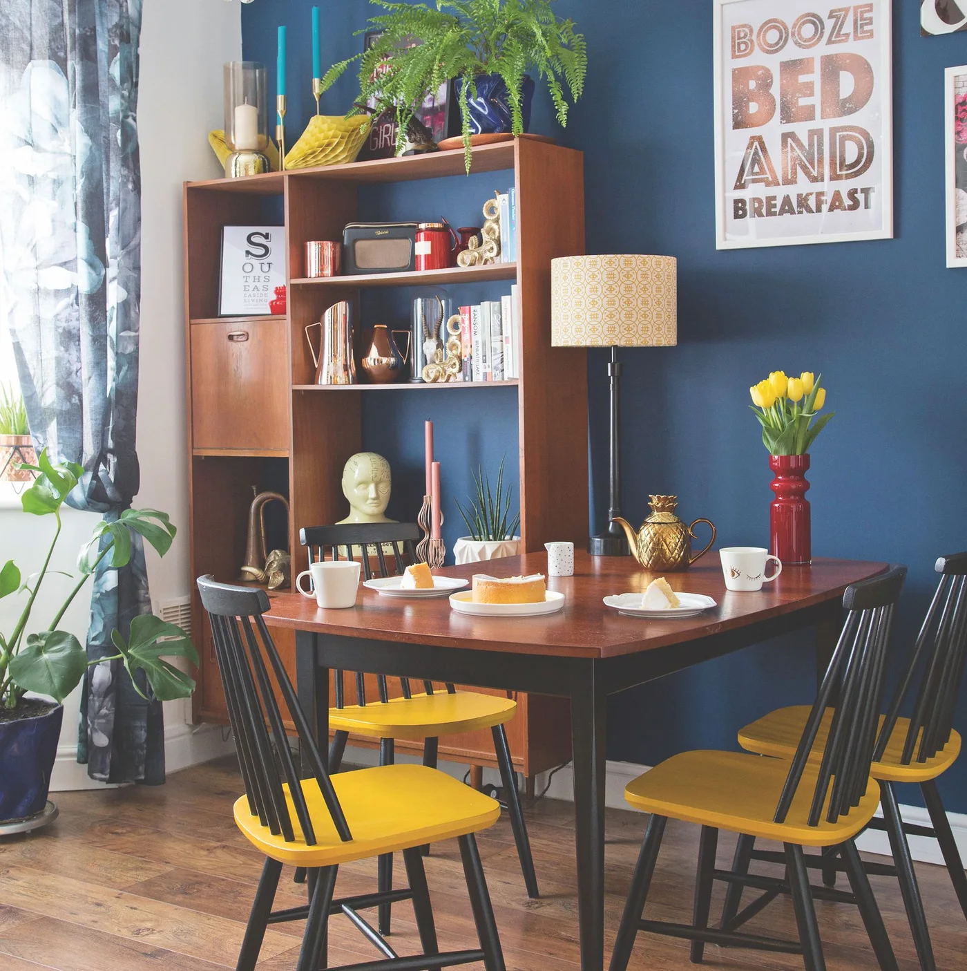 A timber dining table and open cabinet sit against a deep blue wall beside black chairs with yellow seats and a red vase.