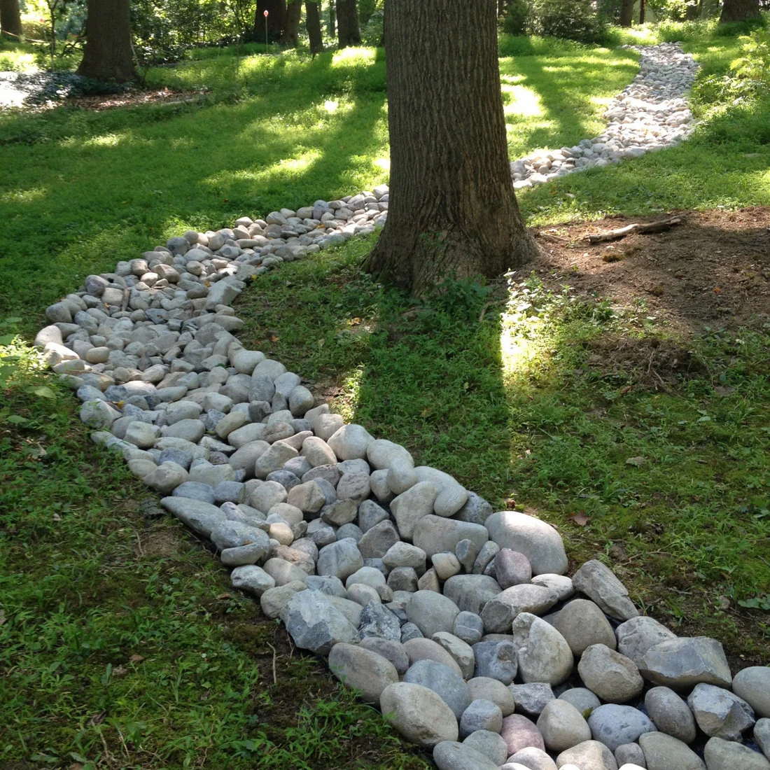 A broad channel of smooth gray and cream stones curves through a lawn around mature trees