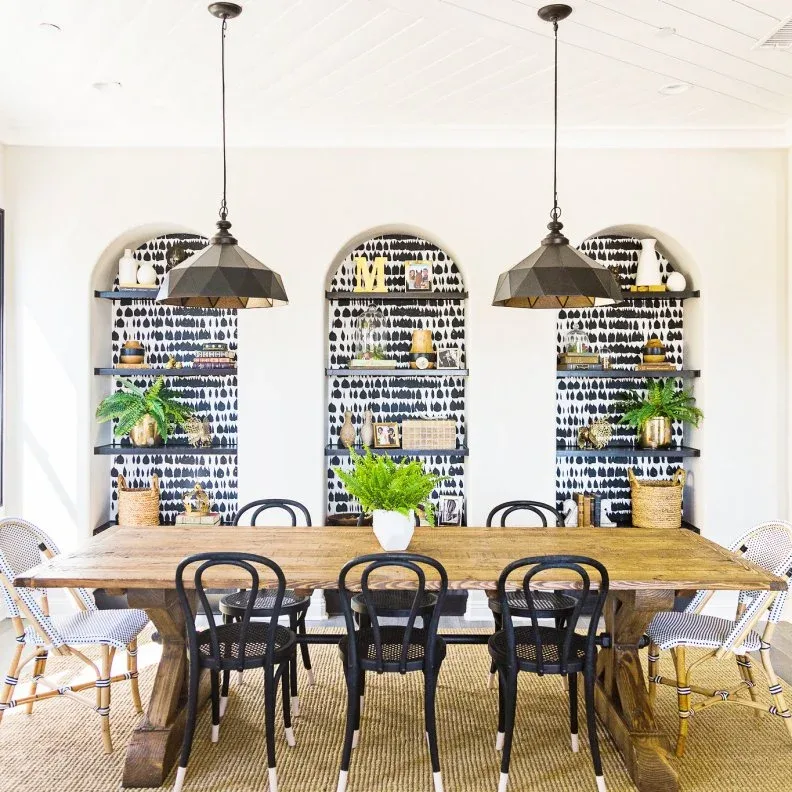 Dining room with three arched wall recesses backed by black-and-white pattern, dark shelves, wooden table, black chairs, pale woven end chairs, and two dark pendants.