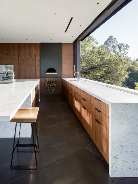 Timber drawers and a pale veined island stand over a dark tile floor beside broad kitchen glazing