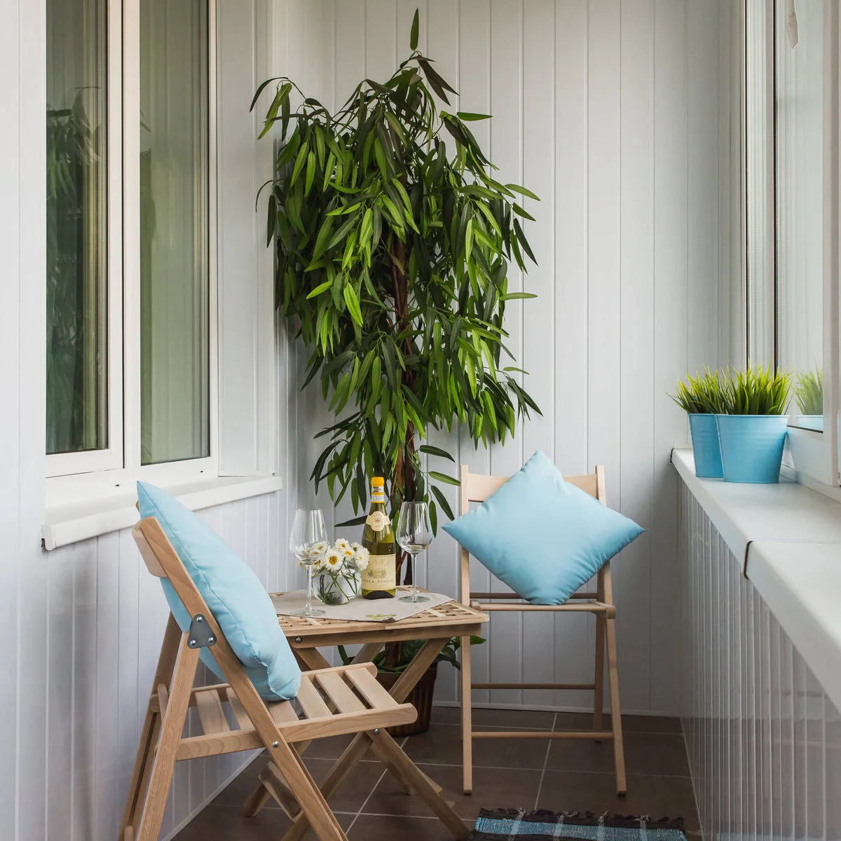 Enclosed narrow balcony with folding wood-colored chairs, small table and blue cushions