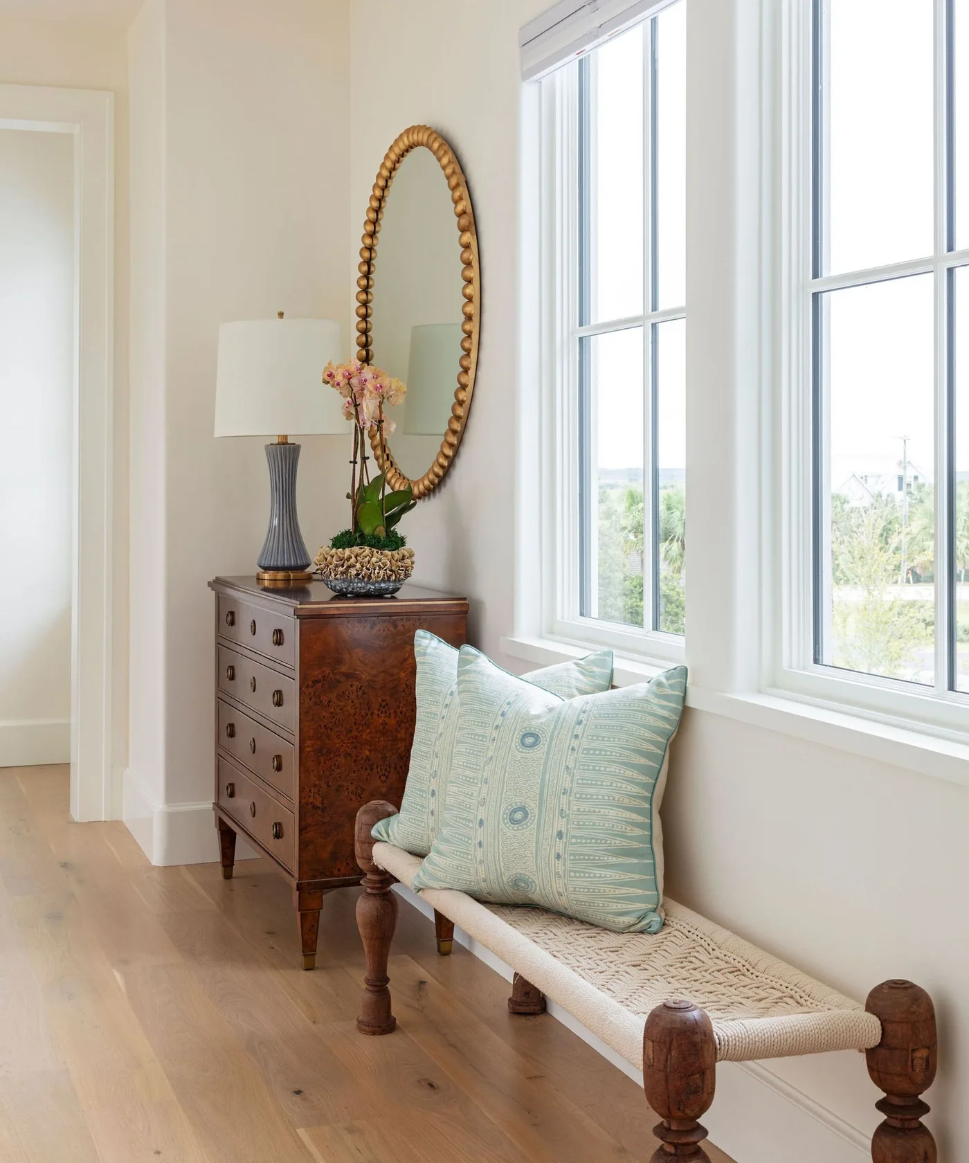 Light entrance with woven wooden bench, pale blue patterned cushions, wooden drawer chest and oval mirror beside large windows.