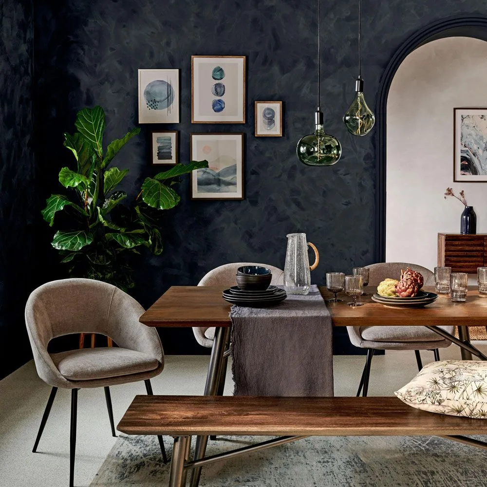 Blue-gray abstract prints in wooden frames with pale mats against a dark textured wall, above a wooden table and wooden bench and gray upholstered chairs beside a green pendant.