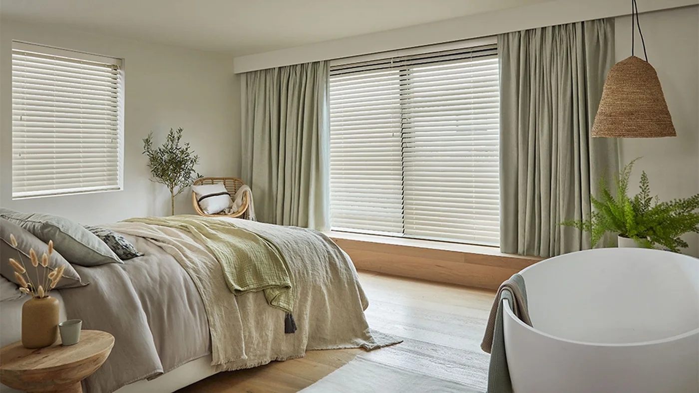 A pale bed with green and cream throws sits beside broad windows with blinds and curtains, a woven pendant and a white bath