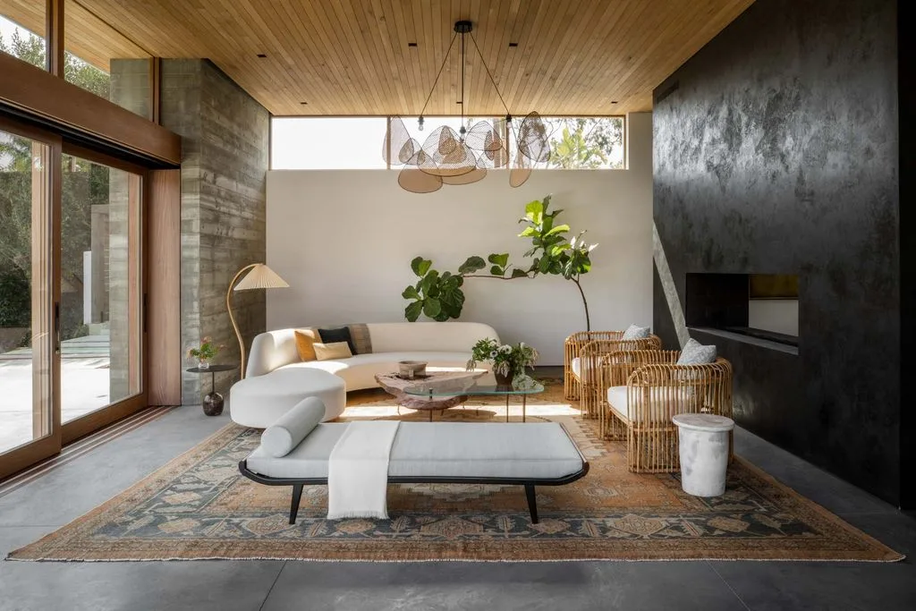 Living room with a wooden ceiling, dark fireplace wall, pale curved sofa, woven chairs and a patterned rug.