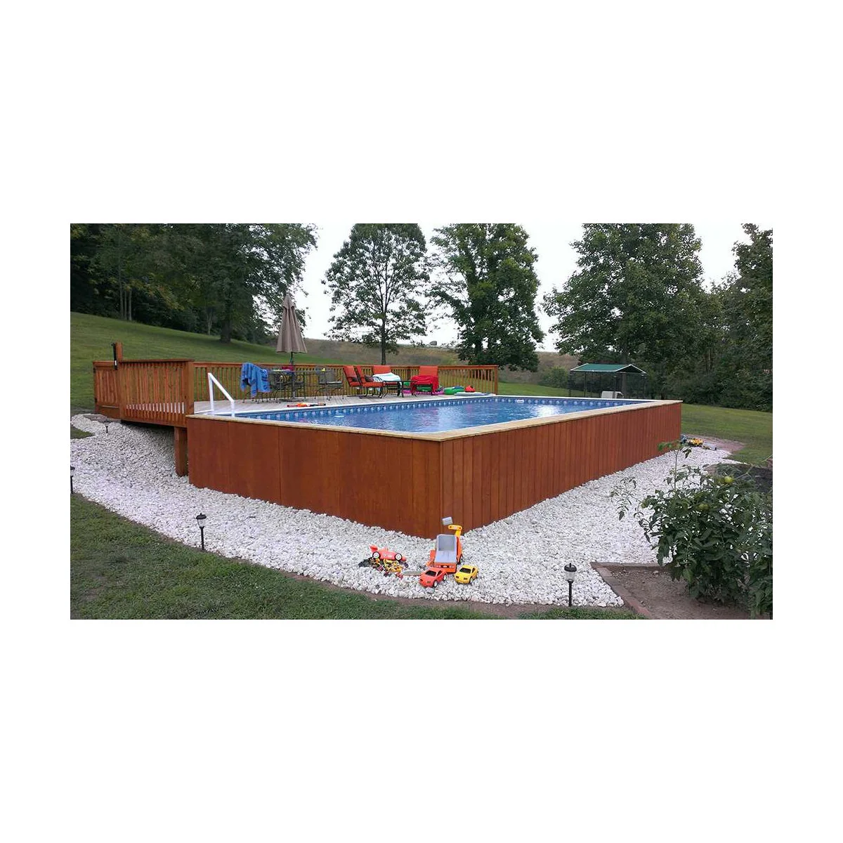 Rectangular above-ground pool with reddish vertical side cladding, a deck at one end and a pale gravel border around the base.