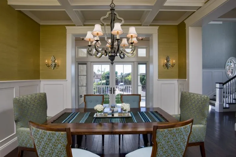 Dining room with ochre upper walls, white paneling and coffered ceiling, green patterned chairs and a dark shaded chandelier