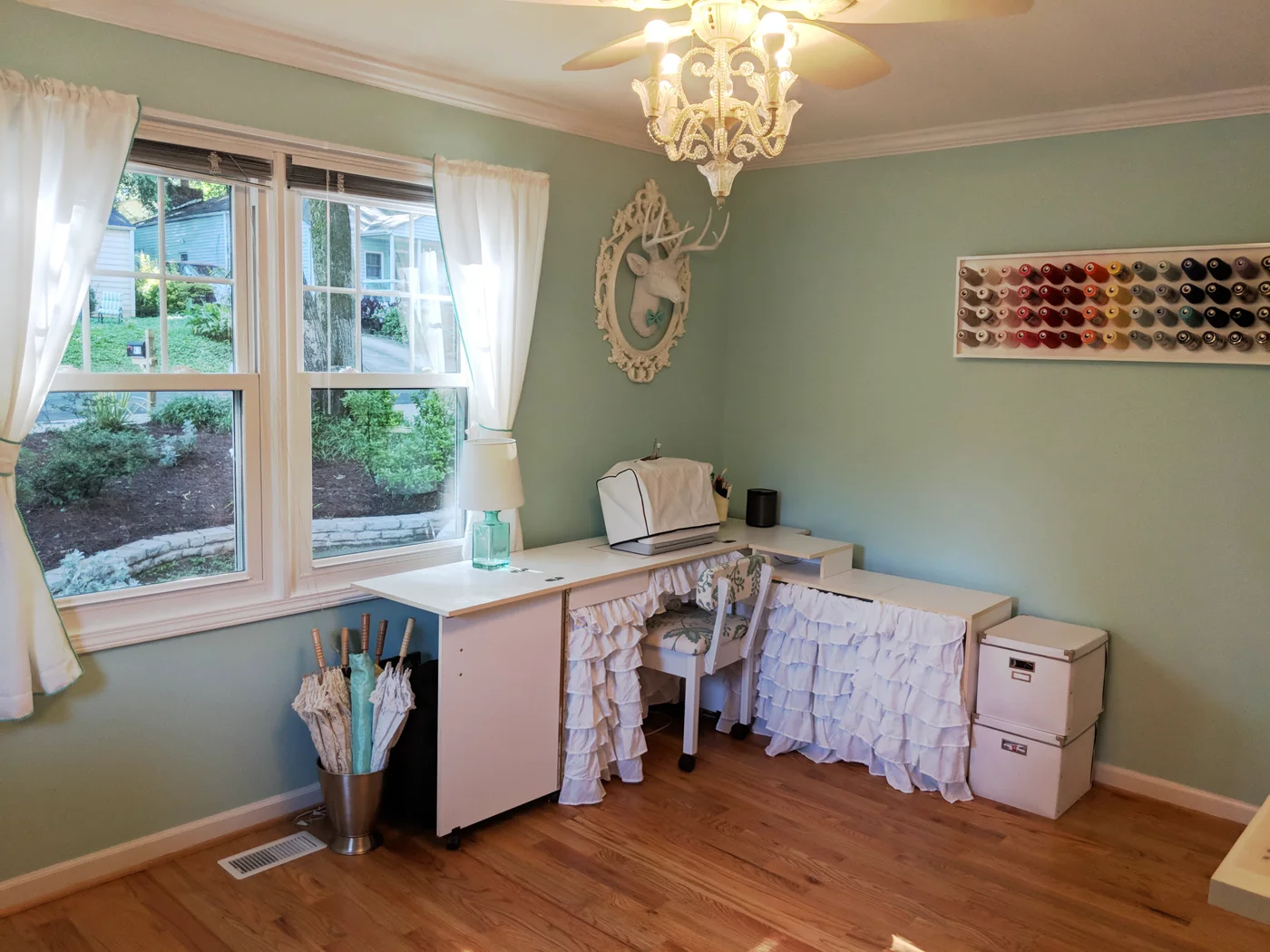 A covered sewing machine sits on a white corner cabinet with a ruffled fabric skirt, a wall thread rack and a small lamp beside the windows.