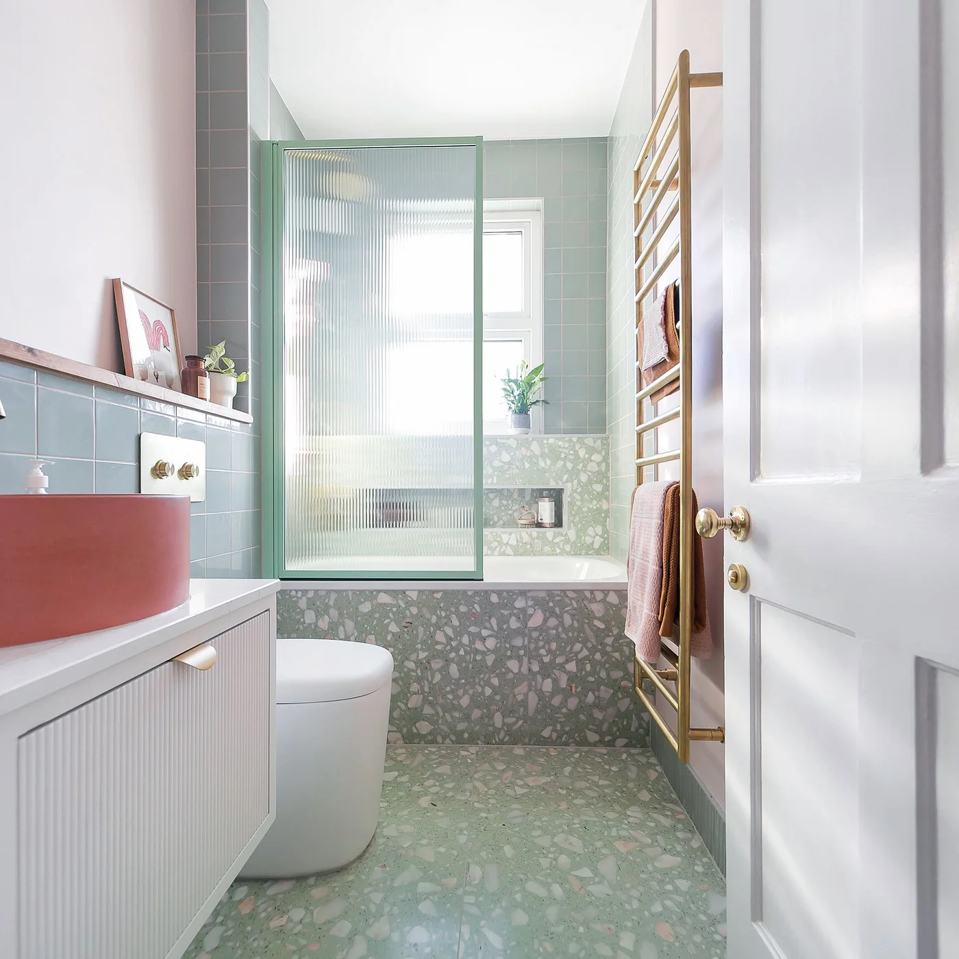 Green terrazzo covers the floor and bath front beside a coral basin, pink walls and a reeded glass screen.