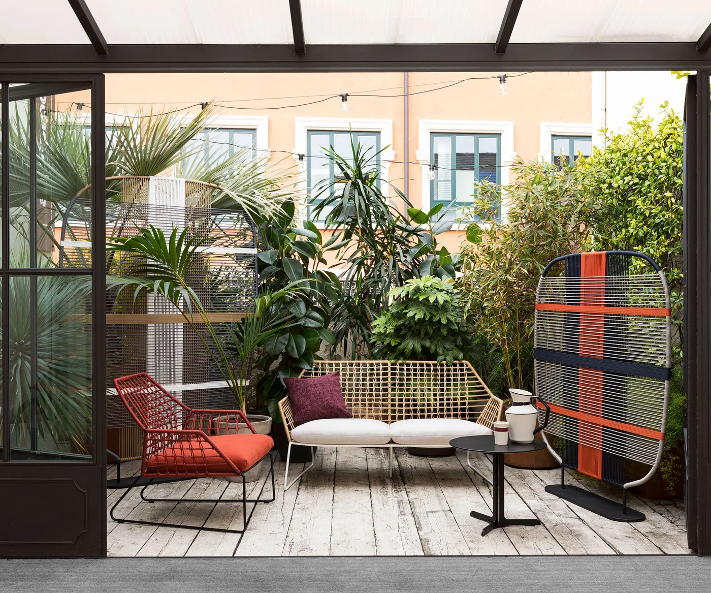 Two patterned freestanding screens and leafy plants surround pale and red outdoor seating