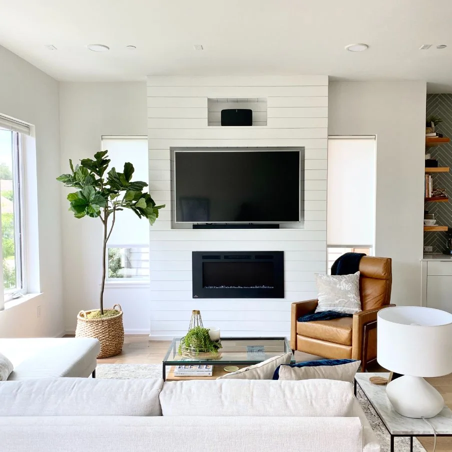 A white horizontal-board feature contains a television, a smaller upper recess and a dark rectangular fireplace, with windows on both sides and pale seating.