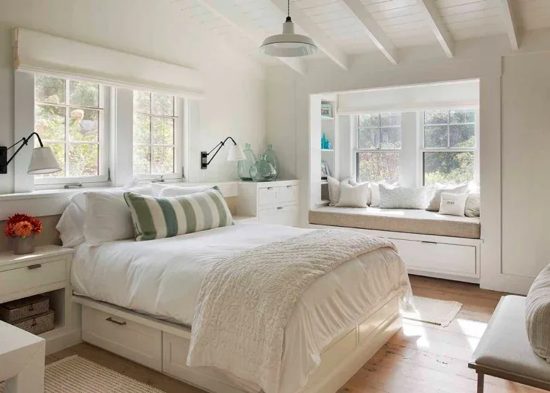 A pale bedroom has exposed ceiling beams, storage drawers under the bed and a cushioned window bench.