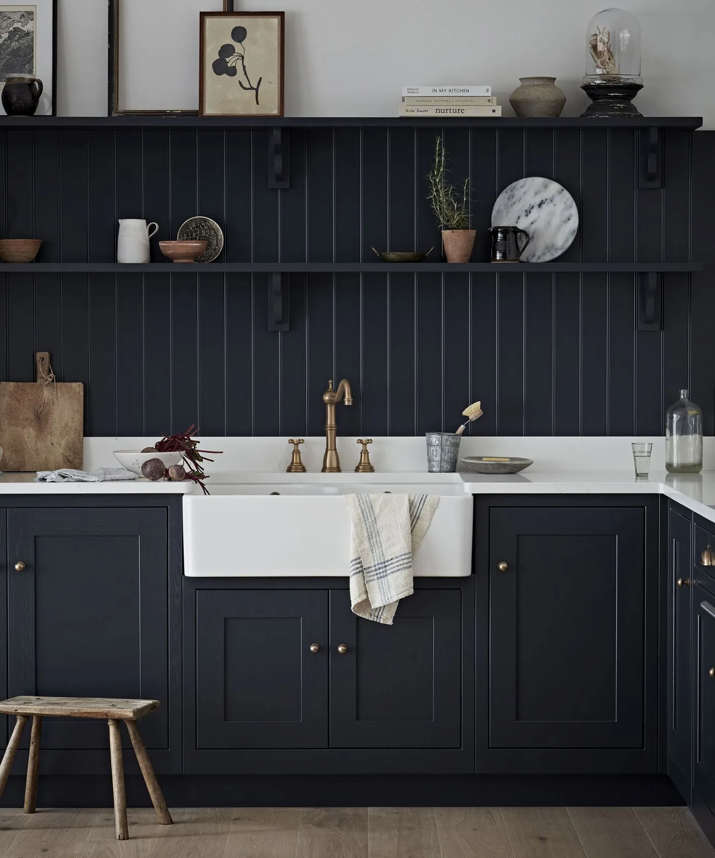 Kitchen with near-black paneled lower cabinets and matching vertical wall paneling, white sink and counter, narrow shelves, warm tap, and wooden stool.