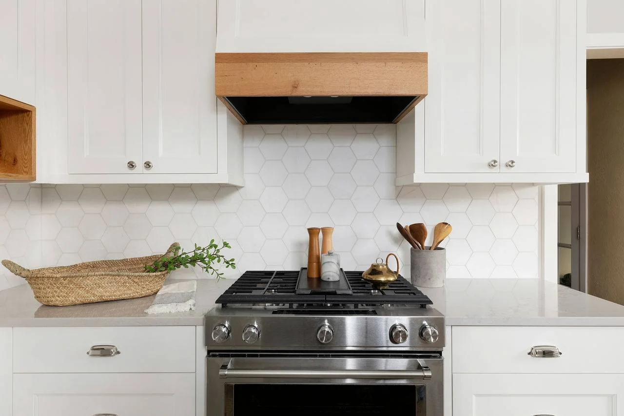 White hexagonal backsplash tiles sit behind a range between simple white cupboards and a timber-edged hood.