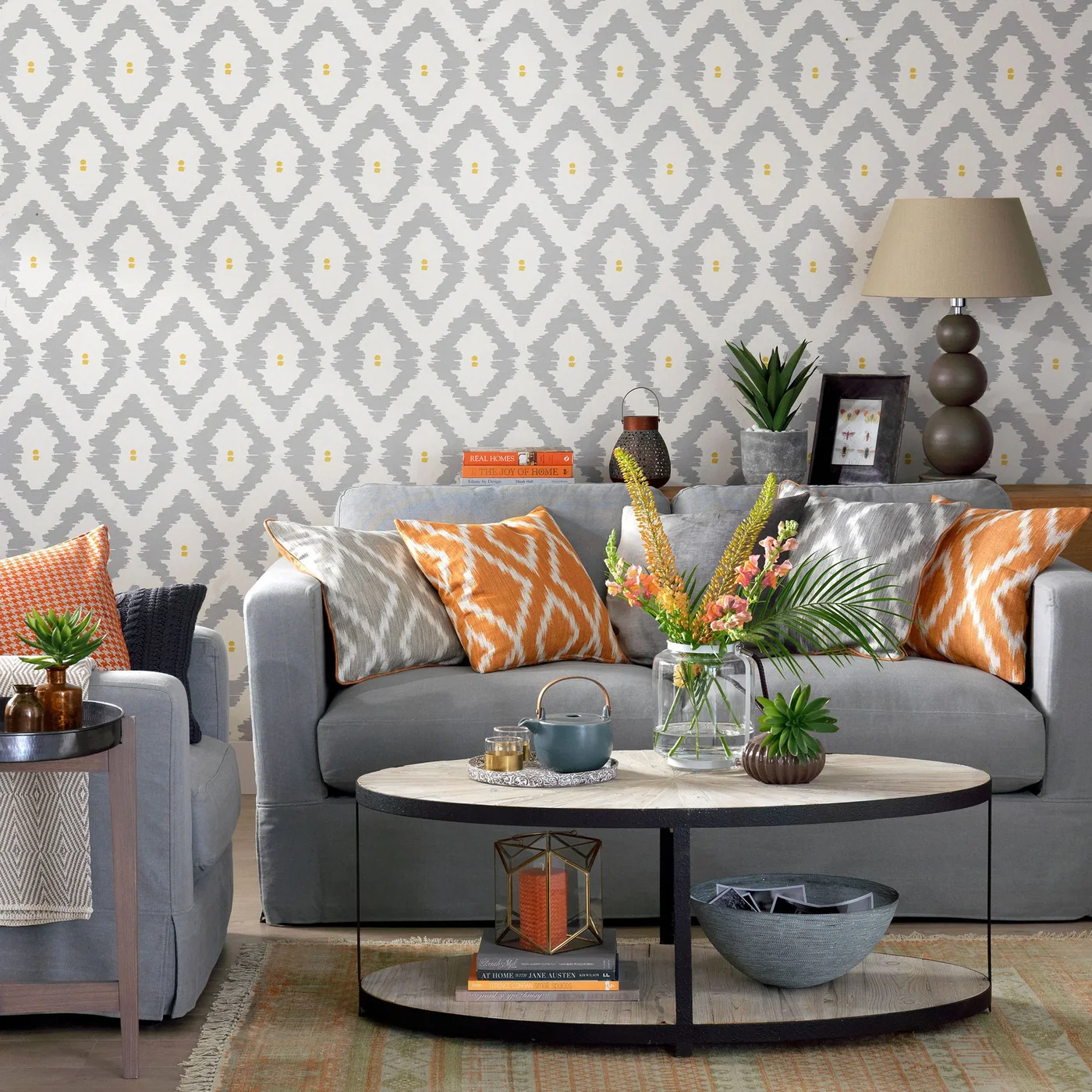 A gray sofa with orange cushions sits against gray-and-white geometric wallpaper beside an oval coffee table