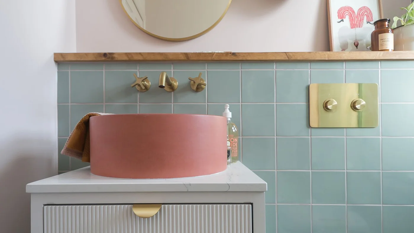 A coral-pink bowl basin stands against mint green square tiles beneath a timber ledge and round mirror.