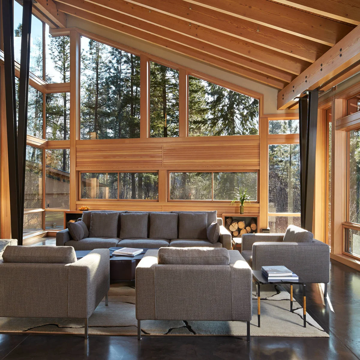 A wood-lined high living room with sloping upper glazing, a gray sofa and three gray chairs arranged around low tables.
