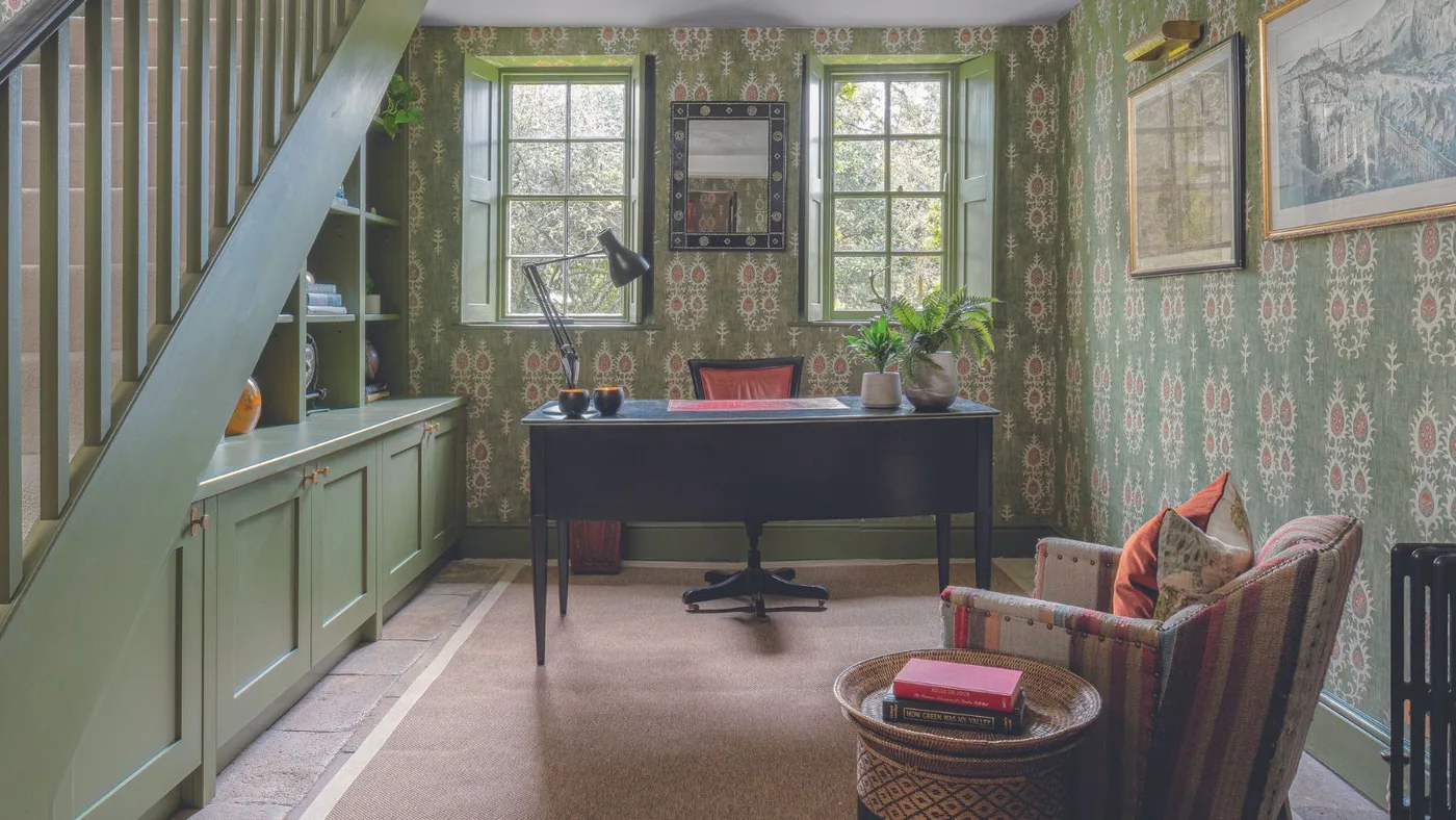 Green patterned office beneath a staircase with a dark desk, framed art and a separate armchair.