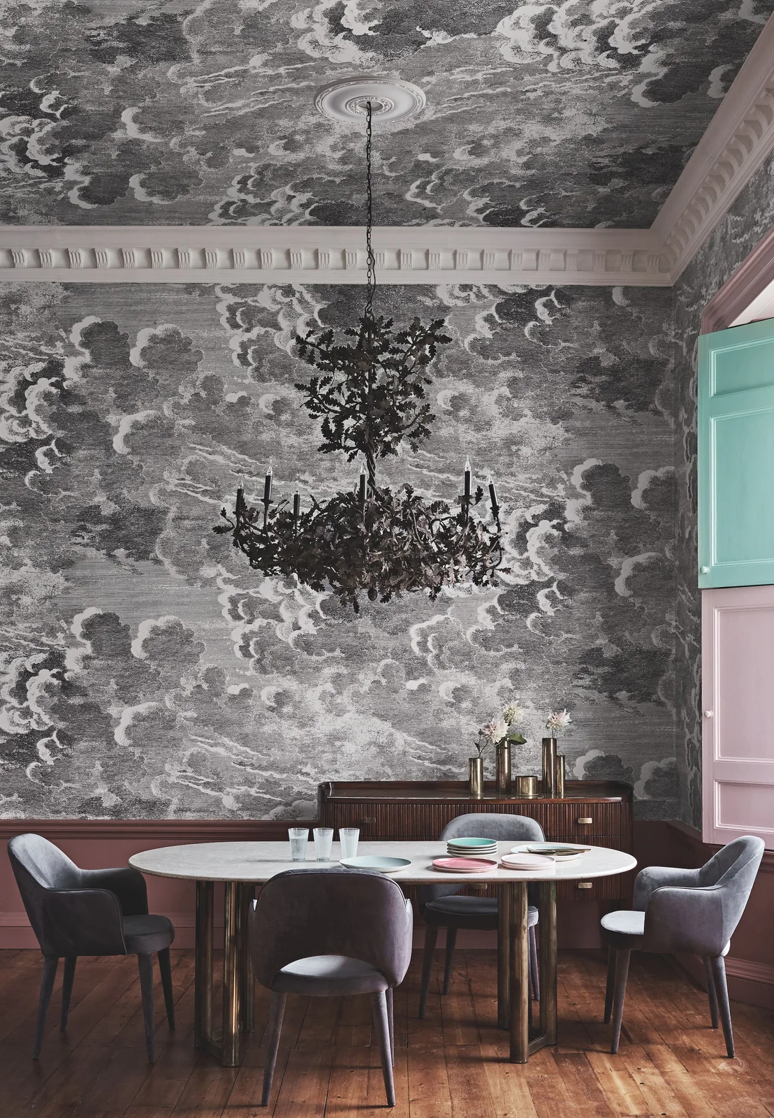 Gray upholstered dining chairs surround a pale oval table beneath a black chandelier and cloud-patterned walls and ceiling
