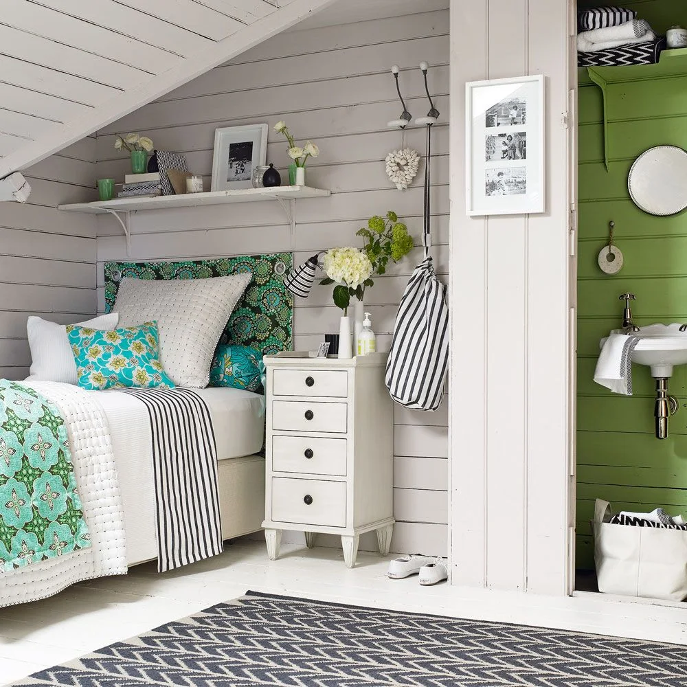 Compact attic bed with green patterned textiles beside a narrow white drawer chest and pale boarded walls.