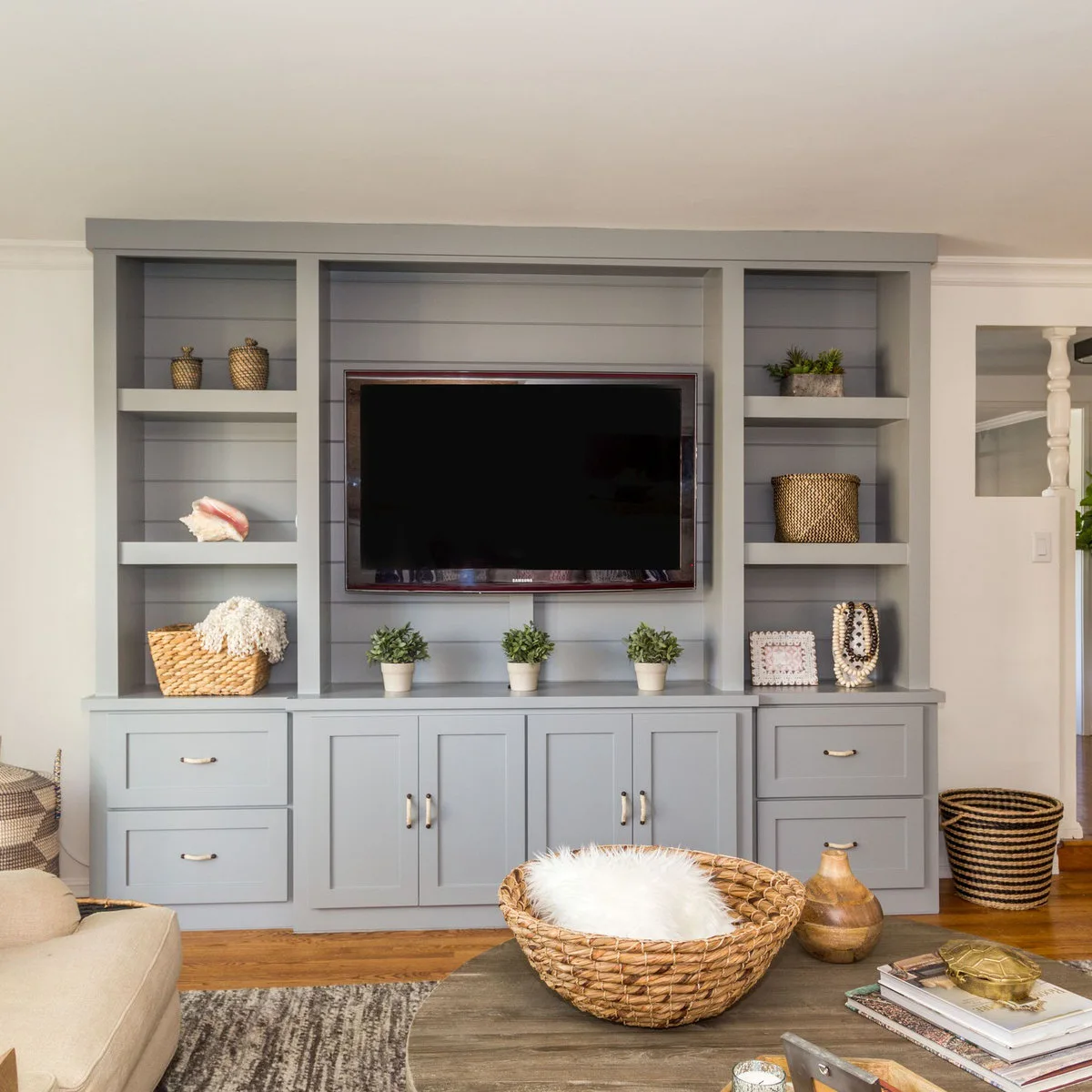 Three small green plants in pale pots sit below a television within gray built-in shelving and lower cabinets.
