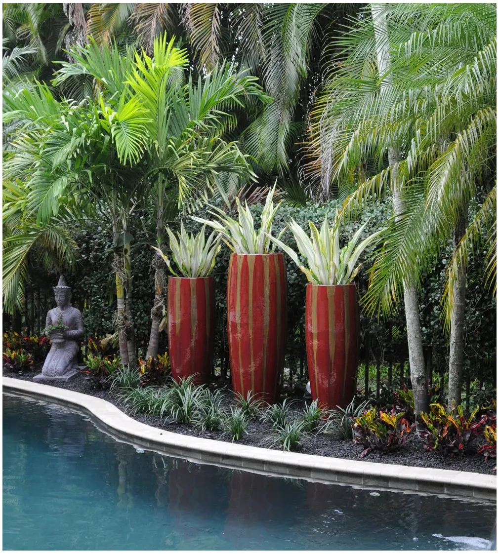 Three tall red-and-gold planters holding upright variegated leaves beside a pool, with palms, low foliage and a seated garden statue.