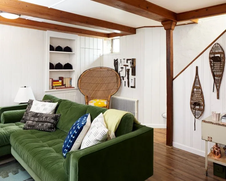 A tufted green sofa sits beneath wooden beams beside a woven high-backed chair and pale boarded walls