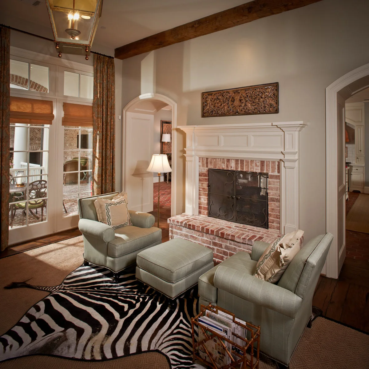 A detailed pale mantel frames red brick beside two muted green chairs, gray walls and tall patterned curtains.