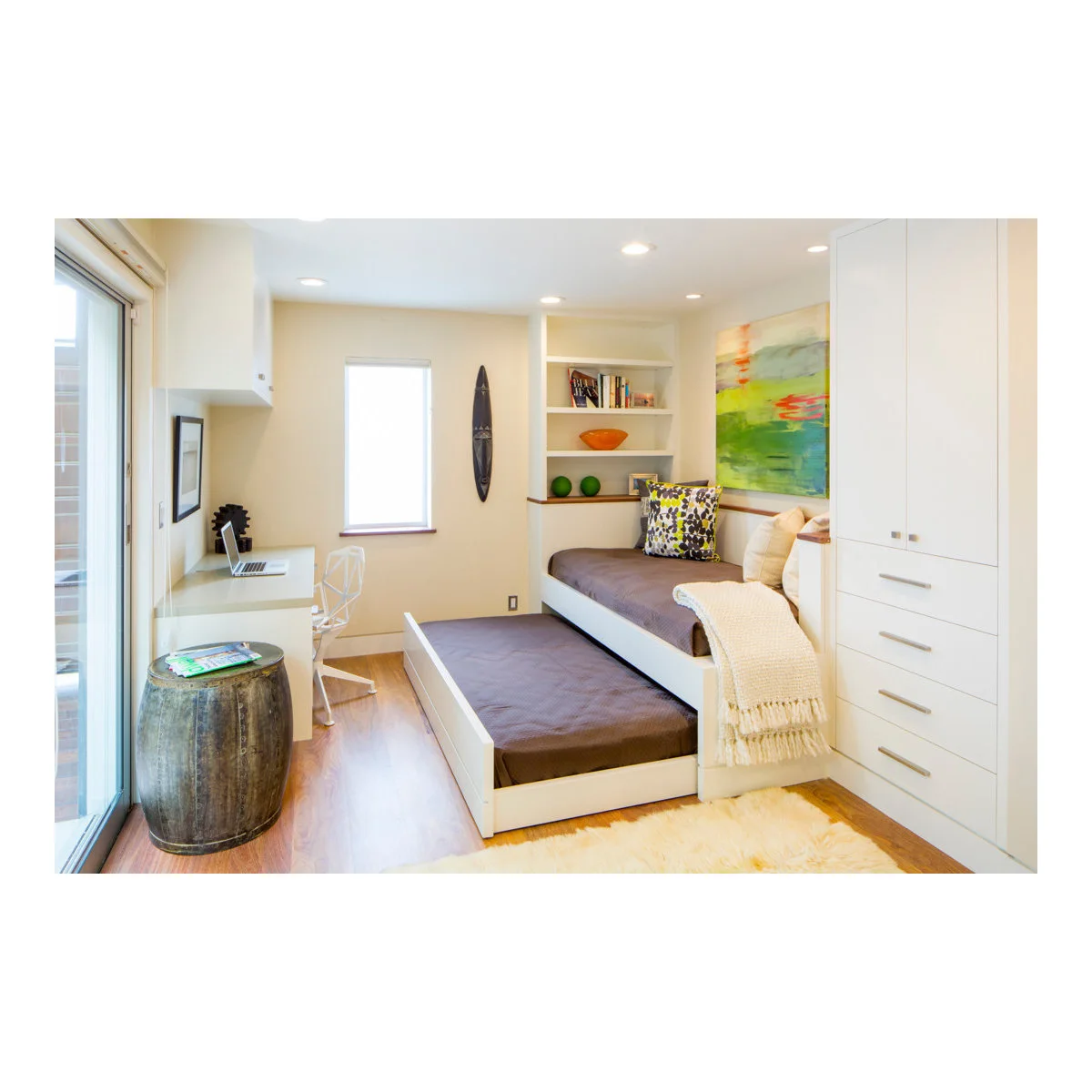 A built-in daybed with a pulled-out lower bed shares a compact room with a desk and tall white storage.
