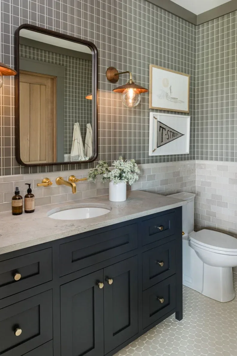 Bathroom with charcoal vanity, pale counter, gray plaid wallpaper, light rectangular lower tiles, rounded dark mirror, warm metal light, and framed prints.