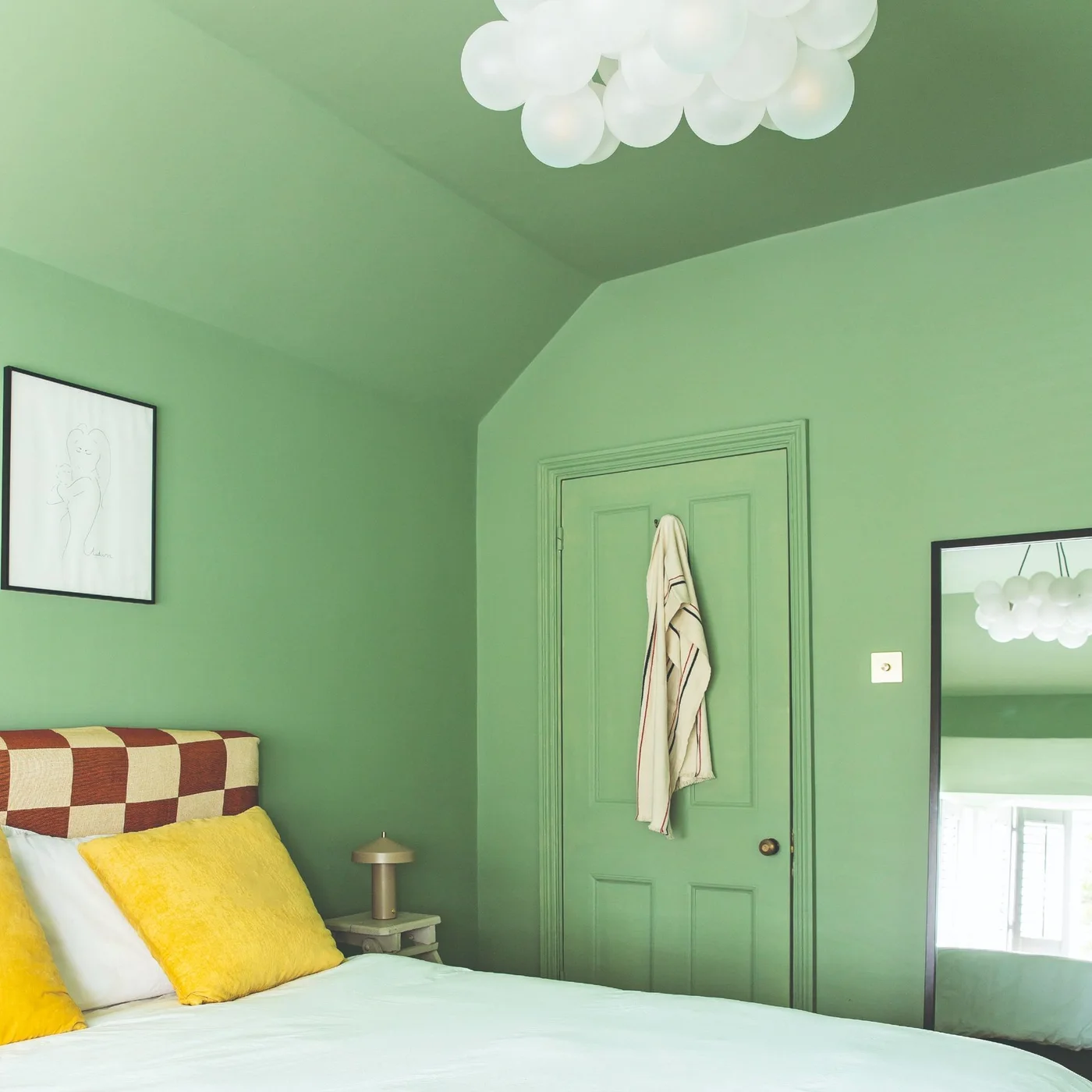 Green walls, trim, door and sloping ceiling surround a bed with a checked headboard and yellow pillows beside a tall dark-framed mirror.