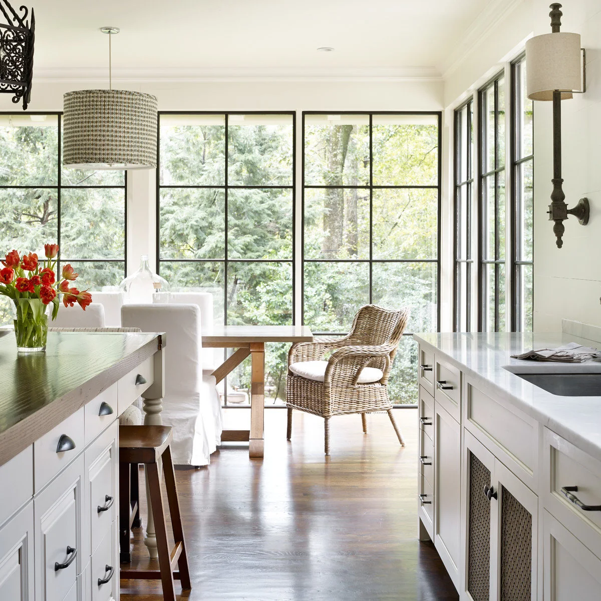 A patterned drum pendant hangs above a breakfast table beside tall windows, while a pale shaded wall light sits near kitchen cabinets.