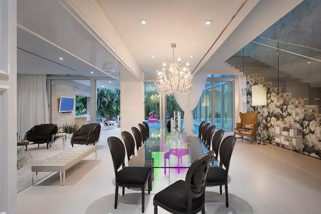 Long clear glass dining table over brightly colored supports surrounded by dark oval-back chairs in an open room