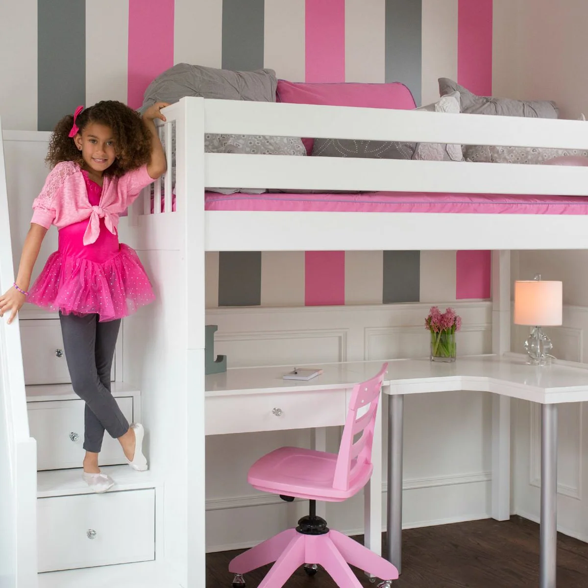 White loft bed against pink and gray striped walls with an L-shaped desk, pink chair and drawer stairs.
