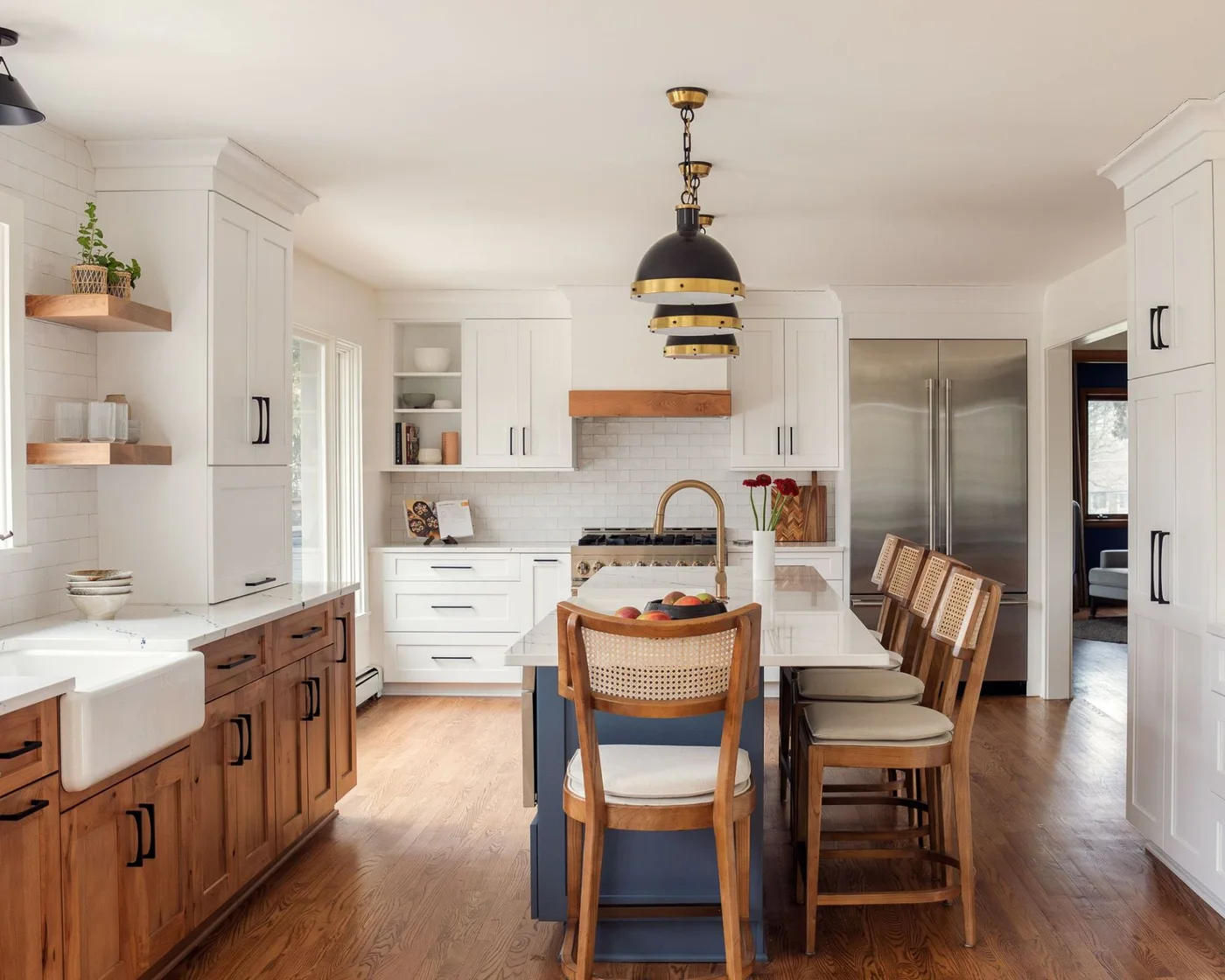 A dark blue island stands beside wood-toned lower cabinets and white upper cupboards beneath dark pendants with woven-backed stools