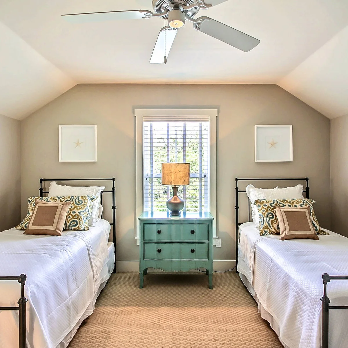 Two black metal beds with white bedding flank a small teal chest beneath a central window