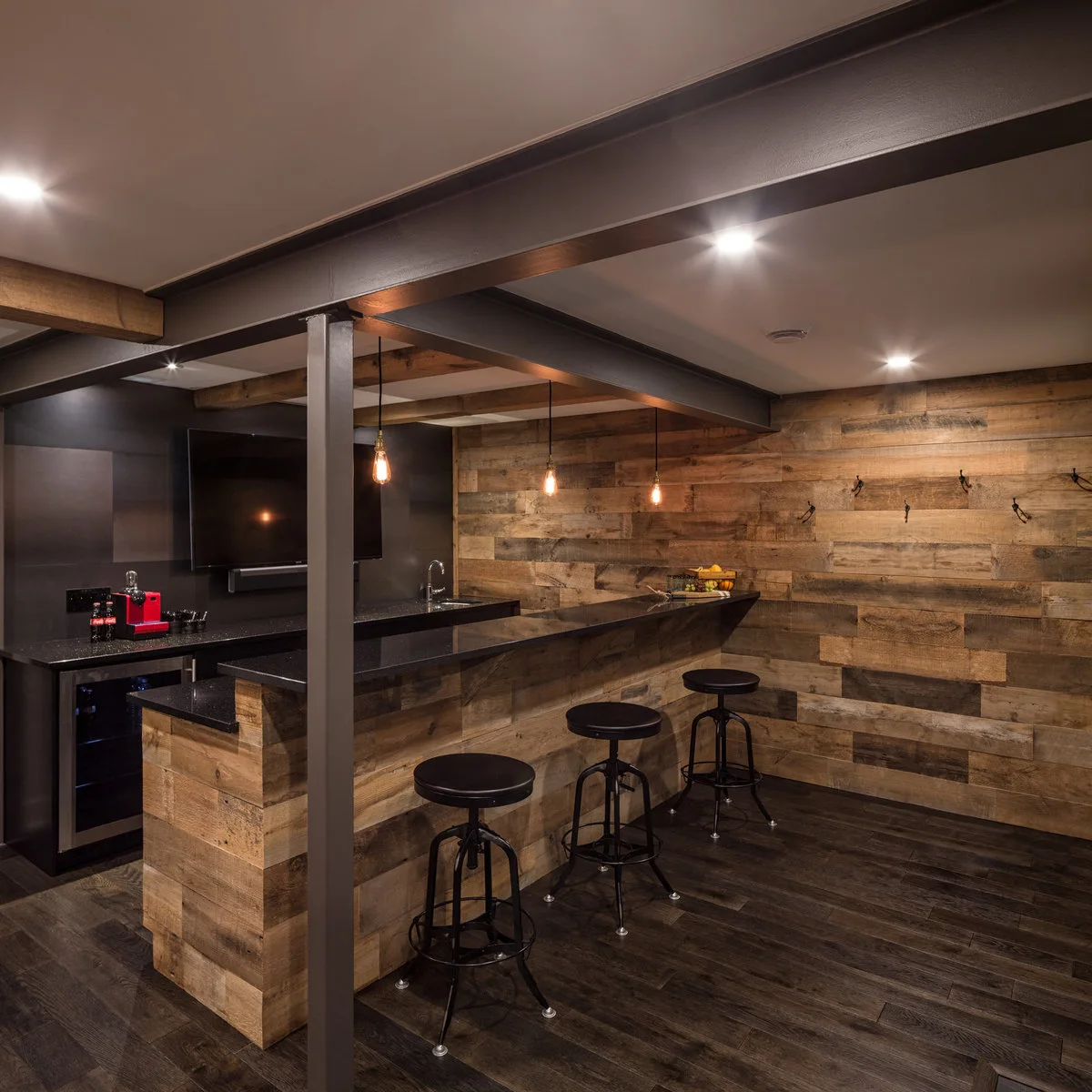 Industrial-style basement bar with wood-colored facing, black stools and exposed dark posts
